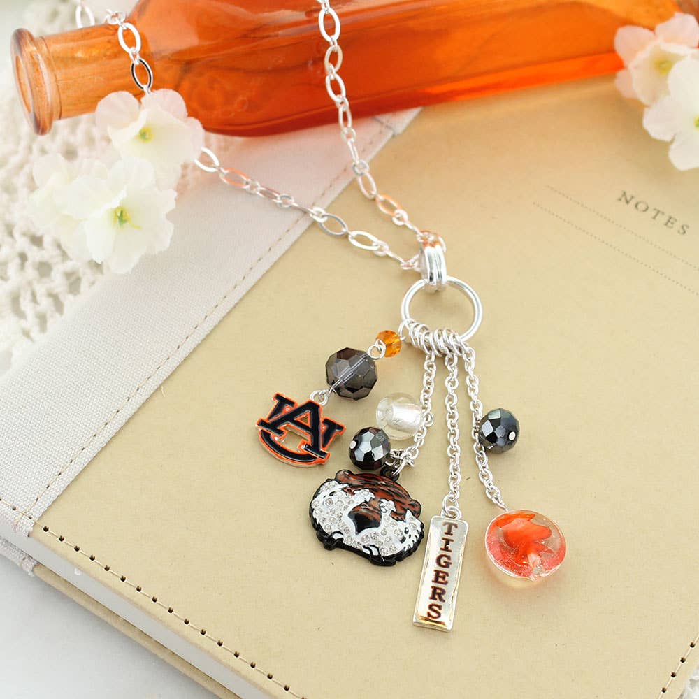 The Auburn Cluster Necklace by Seasons Jewelry, featuring silver-tone Auburn Tigers charms and themed pendants, is displayed on a beige notebook with an orange glass bottle and white flowers in the background.