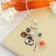 The Auburn Cluster Necklace by Seasons Jewelry, featuring silver-tone Auburn Tigers charms and themed pendants, is displayed on a beige notebook with an orange glass bottle and white flowers in the background.