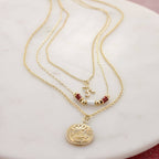 Displayed on a white marble surface, the Alabama Gold & Tube Bead Trio Necklace Set by Seasons Jewelry features three layered gold necklaces—a round pendant, an Om symbol, and red and white bead accents—making it the ideal piece of Bama jewelry for any University of Alabama necklace collection.