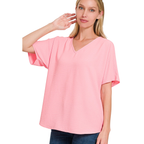 A woman wears the Zenana V-Neck Airflow Shirt Sleeve Top in light pink with blue jeans, posing with one hand near her ear against a white background.