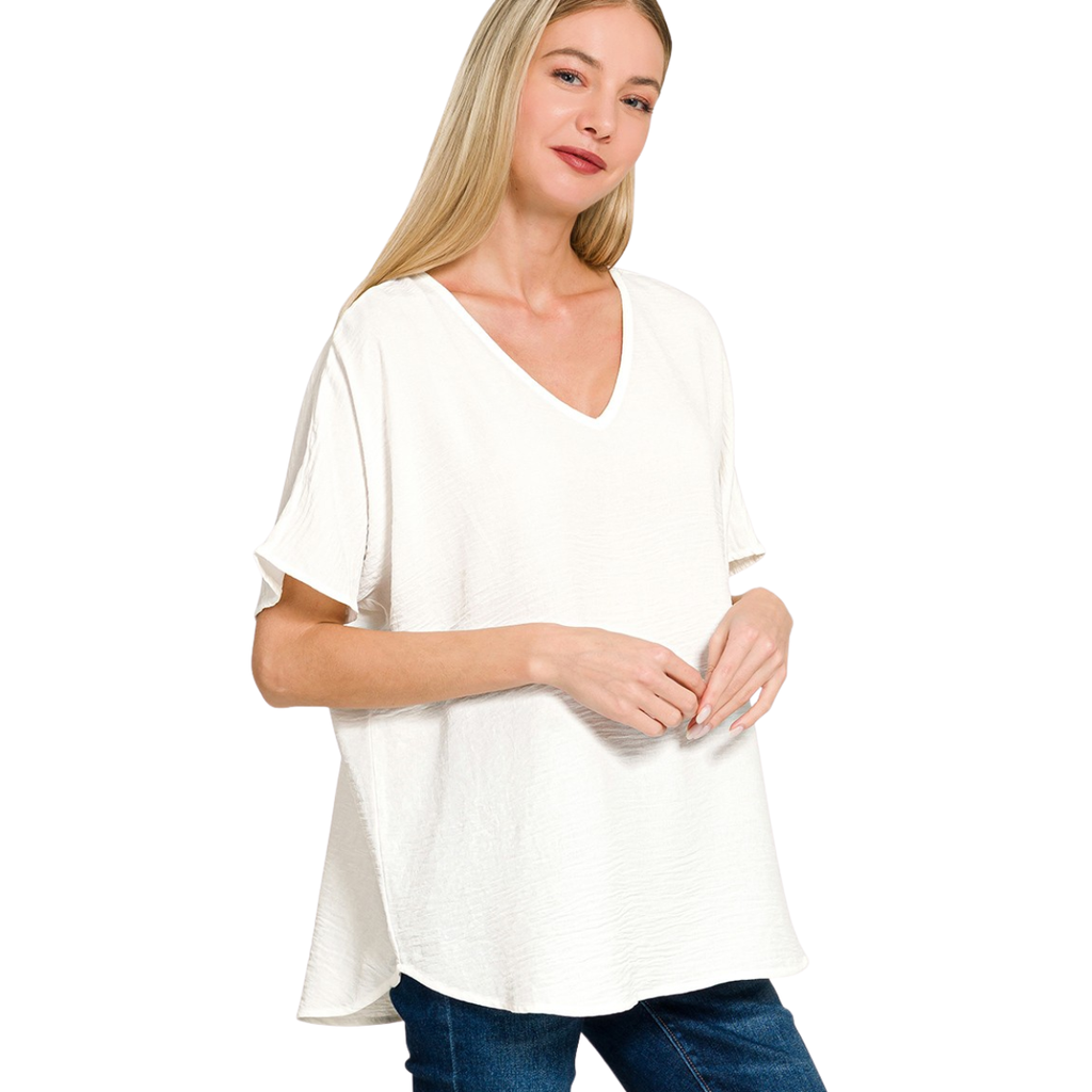 A woman with long blonde hair wears the Zenana V-Neck Airflow Shirt Sleeve Top, a loose-fitting white summer blouse, paired with blue jeans and stands before a plain white background.