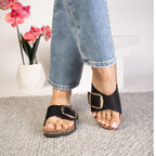 A person wearing blue jeans and CORKY'S FOOTWEAR INC Road Trip Buckle Slide Sandals stands on a gray textured rug, with a white vase of pink flowers in the background.