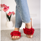 A person in blue jeans and CORKY'S FOOTWEAR INC Wild Child Floral Slide Flat Sandals in Red, with a cushioned cork footbed, stands on a gray rug; pink flowers in a white vase appear in the background.