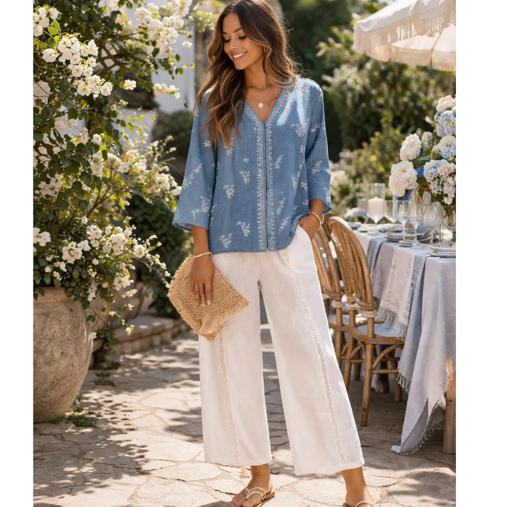A woman stands outdoors by a set dining table, wearing Pine Apparel's Tencel Embroidered Button Front Top in blue, white wide-leg pants, sandals, and holding a woven clutch. She is surrounded by white flowers and greenery.
