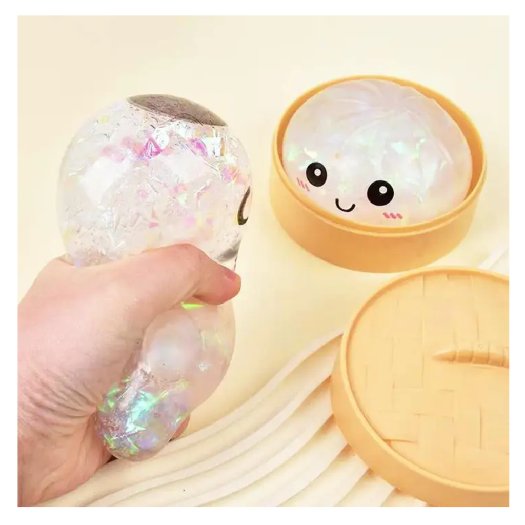 A hand squeezes a clear, iridescent Squishy Dumpling with Steamers by judson & Company beside a round container featuring a smiling dumpling face lid.