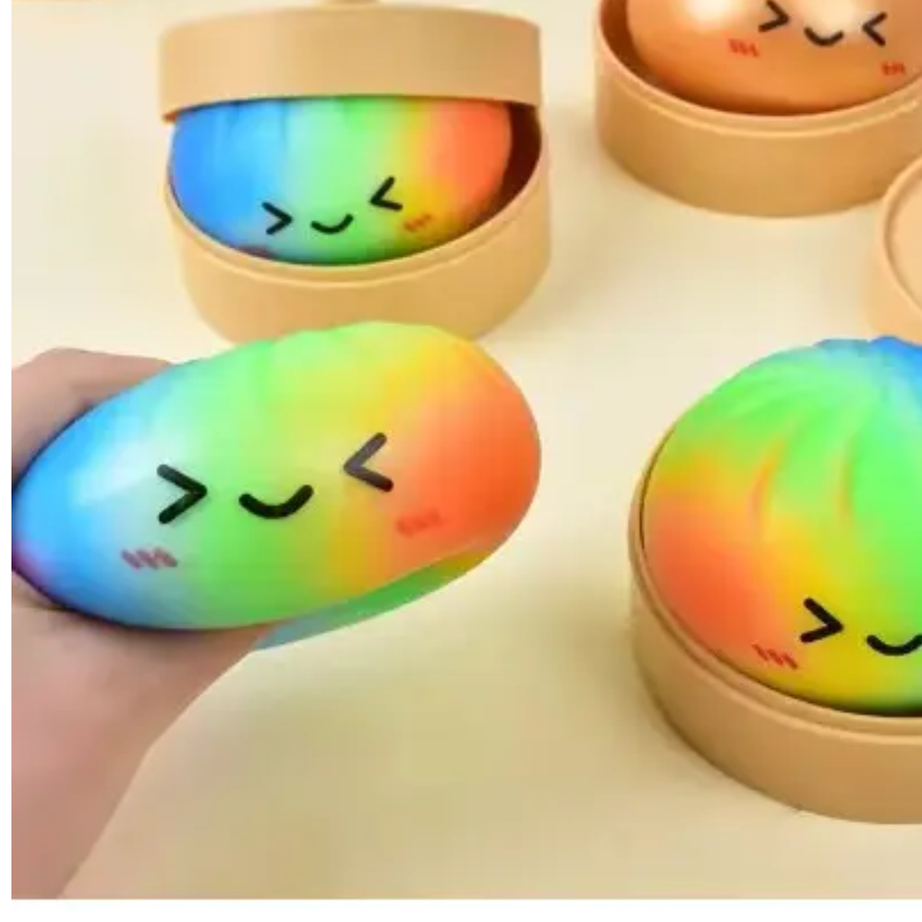 A hand holds a rainbow-colored, winking Squishy Dumpling by judson & Company; these sensory toys come in round tan steamer containers and offer playful stress relief.