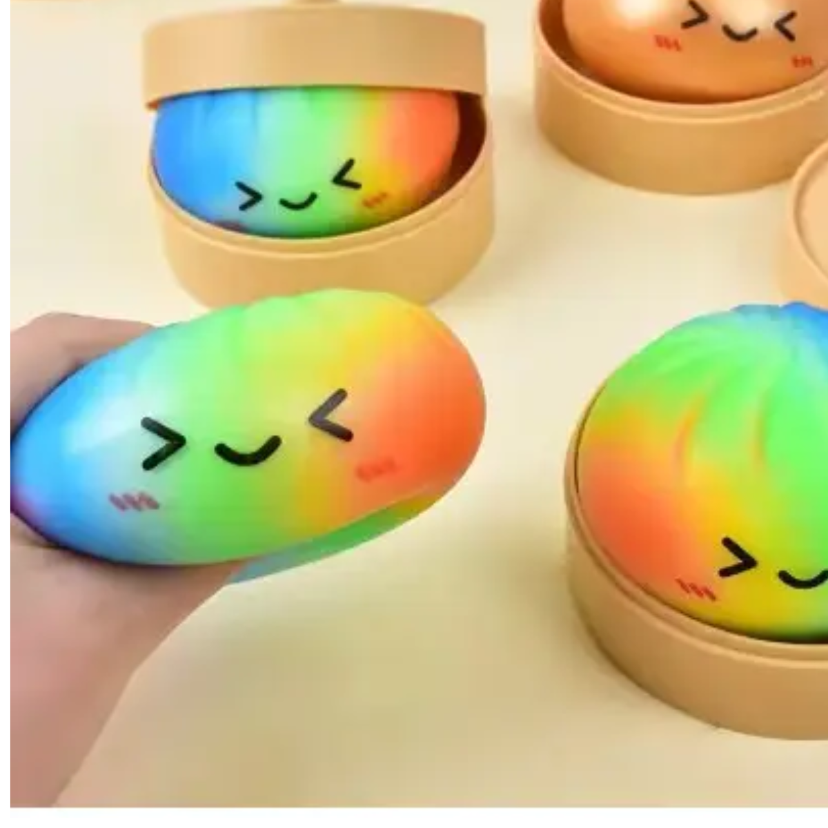 A hand holds a rainbow-colored, winking Squishy Dumpling by judson & Company; these sensory toys come in round tan steamer containers and offer playful stress relief.