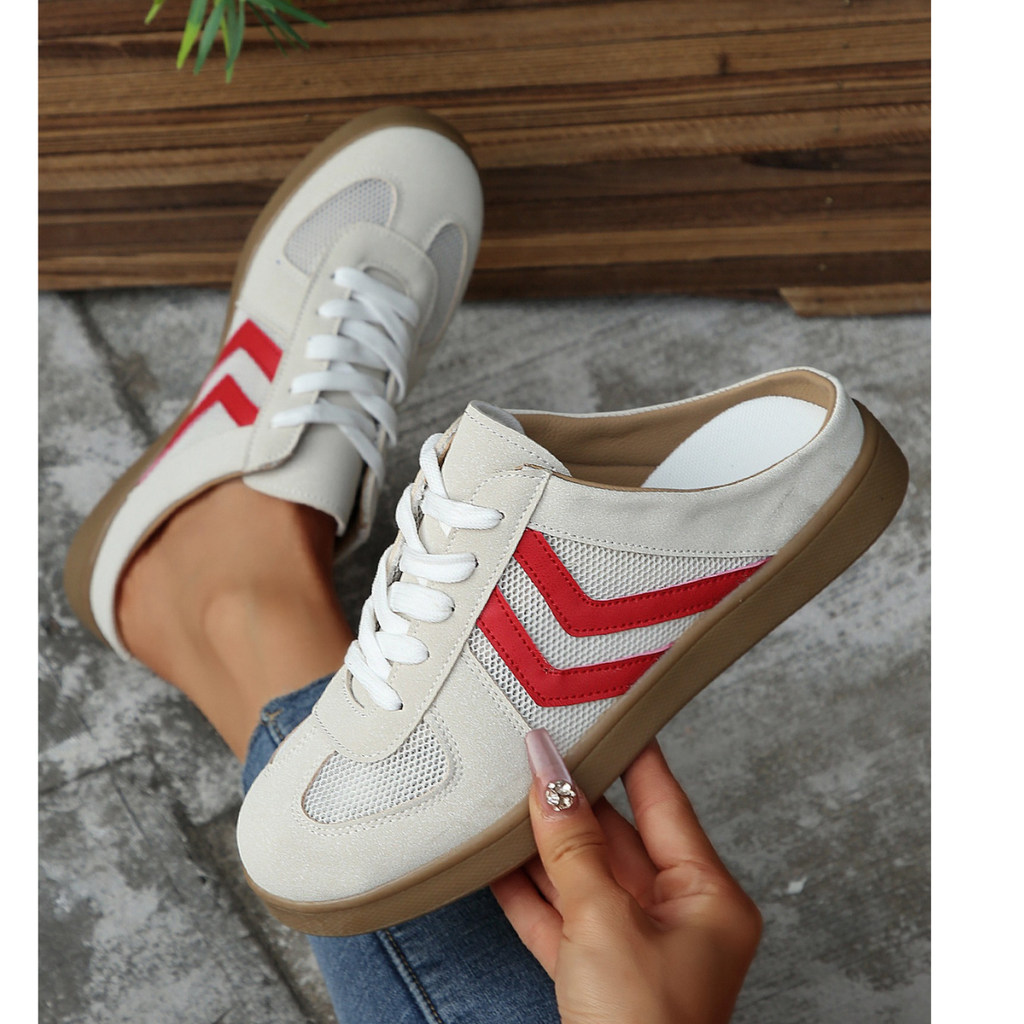 Wearing the FASHION GO Mesh Low Back Slip On Sneakers—featuring white uppers with red chevron stripes, crisp white laces, brown soles, and subtle mesh panels for an athletic touch—a person sits on a concrete floor near wooden planks.