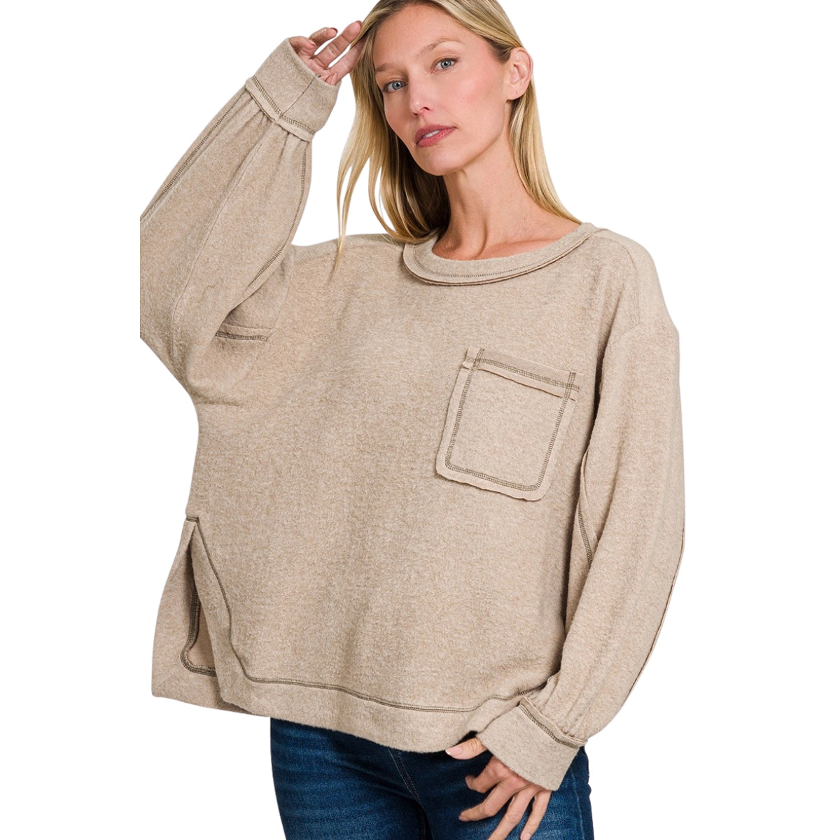 A woman poses against a white background wearing the Fashiongo Brushed Hacci Hi-Low Hem Top in ash mocha, featuring a loose fit, front pocket, and hi-low sweater design, paired with blue jeans.