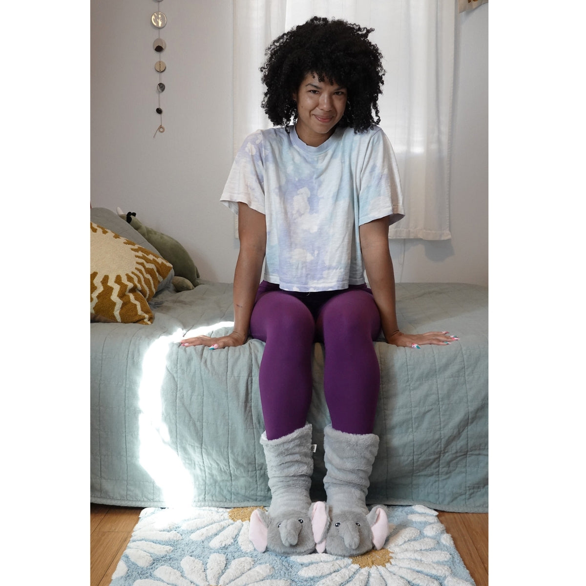 A person sits on a bed in a light-colored room, wearing a tie-dye shirt, purple leggings, and FASHION GO Elephant Cozy Slipper Socks with non-skid soles, as sunlight streams in from the left. Main image