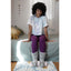 A person sits on a bed in a light-colored room, wearing a tie-dye shirt, purple leggings, and FASHION GO Elephant Cozy Slipper Socks with non-skid soles, as sunlight streams in from the left.