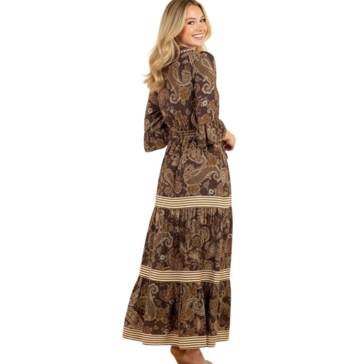 A woman stands barefoot, looking over her shoulder in the FASHION GO Brown Boho Print Paisley Dress, a long-sleeved maxi style with a tiered skirt, beige stripe accents, and a subtle split V-neckline.