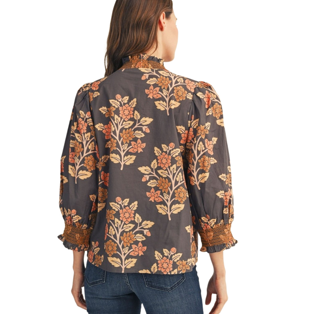 A woman is seen from behind wearing the FASHION GO Embroidered Smocking Detail Floral Top, a long-sleeved dark blouse with an orange floral print, ruffled cuffs, and embroidered smocking, paired with blue jeans.