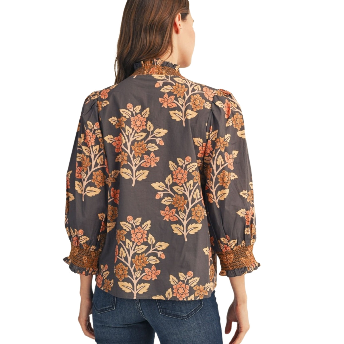 A woman is seen from behind wearing the FASHION GO Embroidered Smocking Detail Floral Top, a long-sleeved dark blouse with an orange floral print, ruffled cuffs, and embroidered smocking, paired with blue jeans.