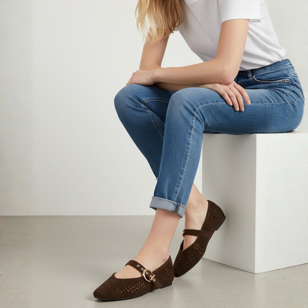 A person in a white shirt and blue jeans sits with crossed legs on a white block, wearing Top Guy Perforated Upper Ballet Flat Mary Jane shoes.