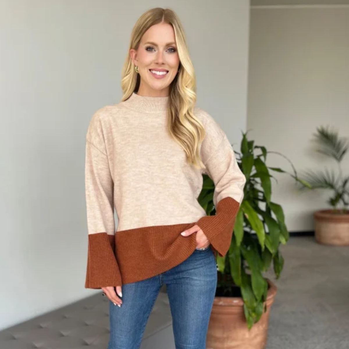 A woman smiles at the camera indoors, wearing the Carole Christian Color Block Sweater with blue jeans. Potted plants and neutral-colored walls are in the background.