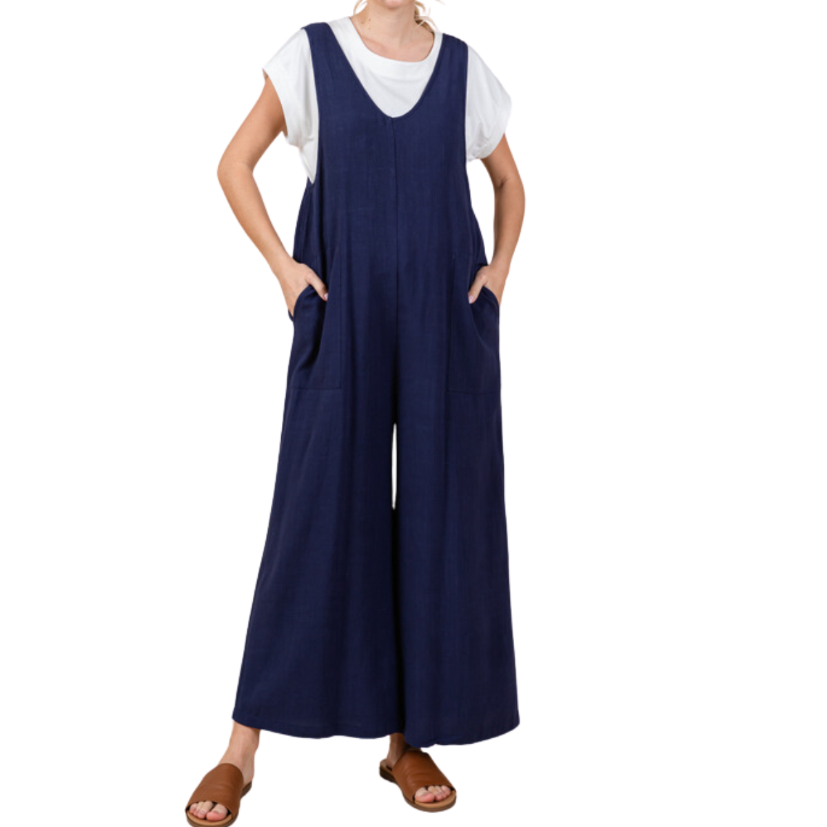 A person is wearing a white t-shirt underneath a loose-fitting, dark blue Jumpsuit by Jodifl with hands in pockets. They are also wearing brown open-toed sandals. The head and upper shoulders are cropped out of the image.