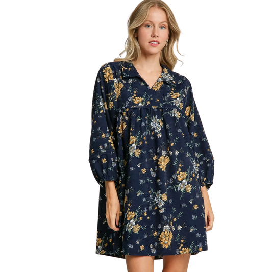 A woman wearing the Umgee Floral Print Corduroy Short Dress, a loose-fitting navy A-line dress with yellow and green floral patterns, stands against a white background.