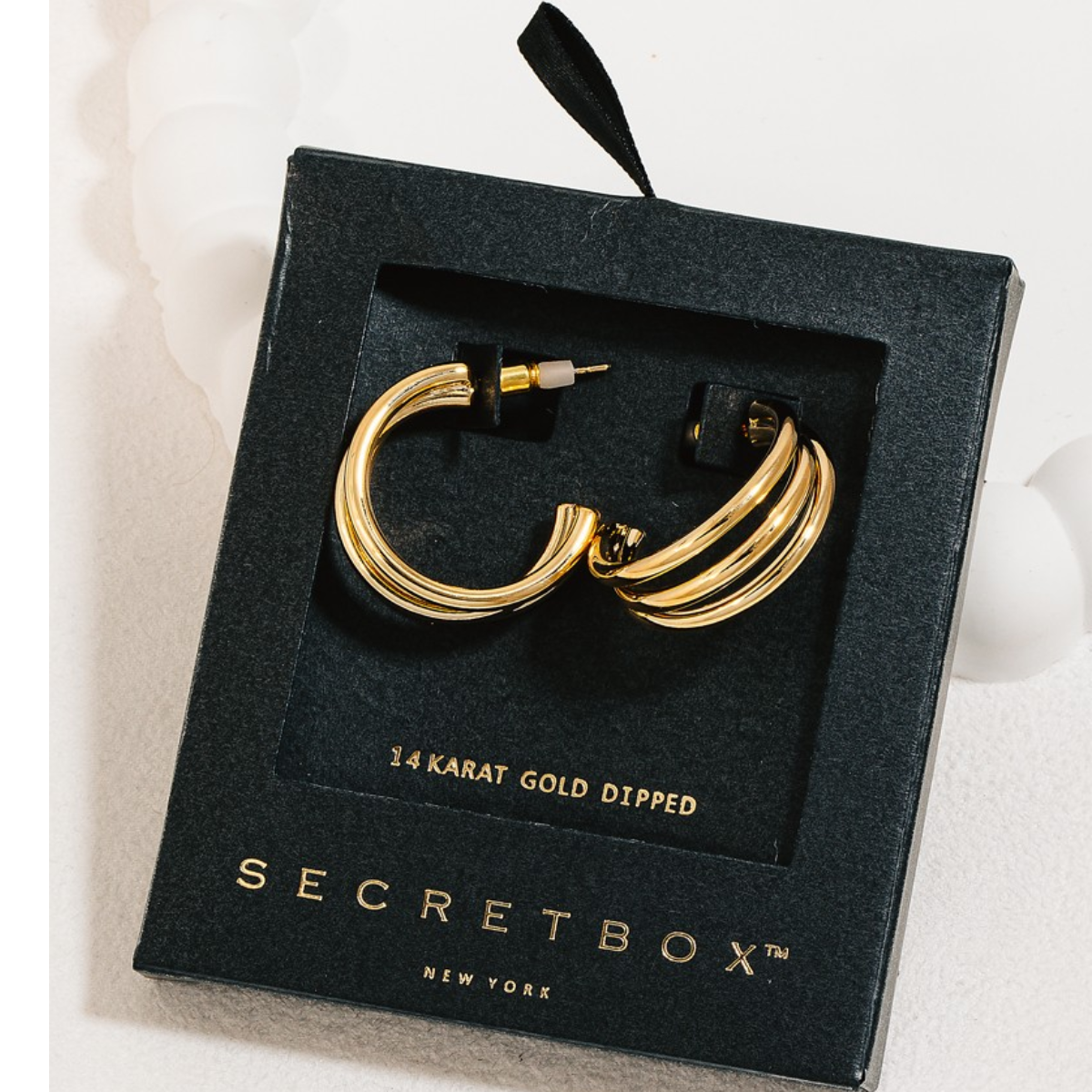 A pair of Secret Box Gold Dipped Layered Tube Hoop Earrings in Gold, 14 karat gold dipped hoop earrings rests inside a sleek black box adorned with the brand name "FASHION GO.