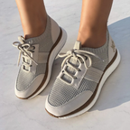 A person wears Speed Sneakers in Mist by OTBT from Consolidated Shoe Co—light gray women's sneakers with white laces and a recycled rubber sole—standing on a light-colored surface.