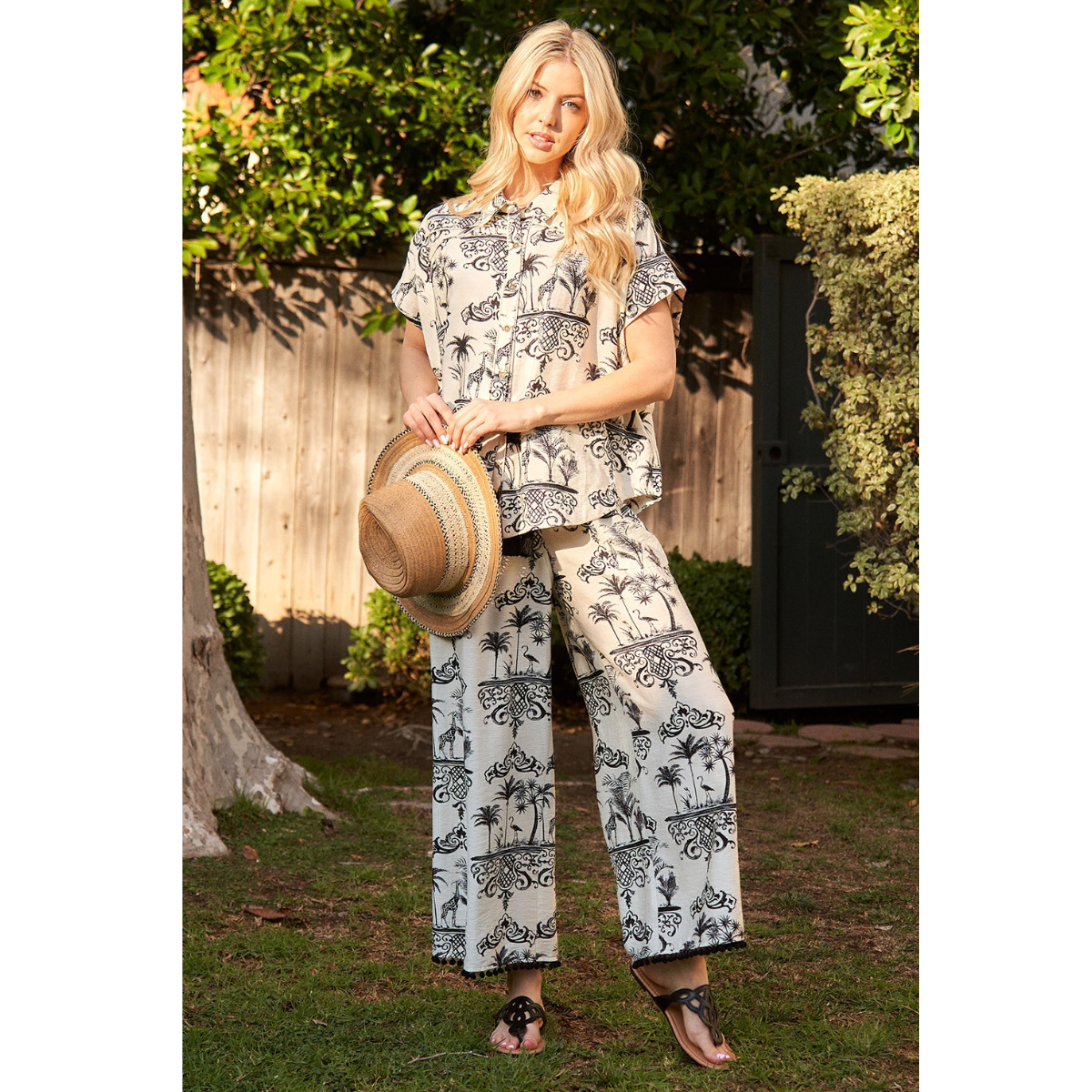 A woman stands outdoors in a yard wearing the FASHION GO Toile Print 2 piece Set—matching short-sleeve top and relaxed fit pants with a tassel tie—holding a straw hat, with greenery and a wooden fence behind her. Main image
