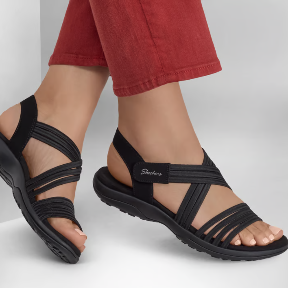 Person wearing black SKECHERS Reggae Slim Sunnyside sandals with a cushioned footbed and red cropped pants, standing on a light gray surface.