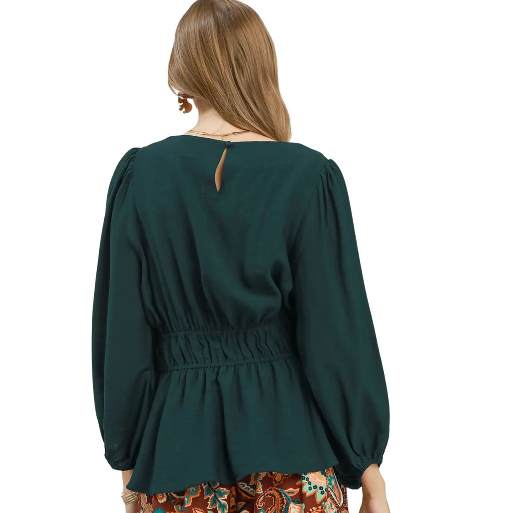 A woman faces away from the camera wearing the Umgee Smocked Waist Long Sleeve Peplum Blouse in dark green paired with patterned pants.
