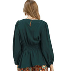 A woman faces away from the camera wearing the Umgee Smocked Waist Long Sleeve Peplum Blouse in dark green paired with patterned pants.