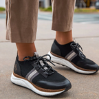 A person in beige pants stands outdoors on concrete, wearing the Speed Sneakers in Black by OTBT from Consolidated Shoe Co, featuring gray stripes and white recycled rubber soles for added bounce back technology.