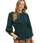 A woman models the Umgee Smocked Waist Long Sleeve Peplum Blouse in dark green, featuring a gathered waist, paired with a patterned brown skirt, as she poses and smiles against a white background.
