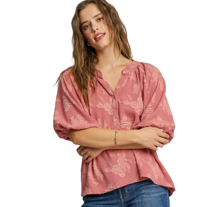A woman with long brown hair models the Umgee Textured Floral Puff Sleeve Blouse in rose, paired with blue jeans, standing against a white background.