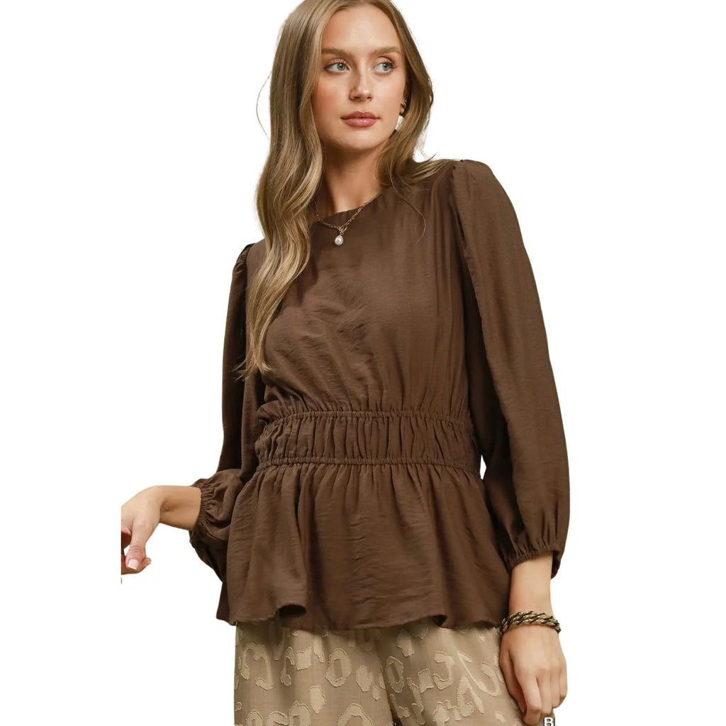 A woman wears the Umgee Smocked Waist Long Sleeve Peplum Blouse paired with beige patterned pants, standing against a white background.