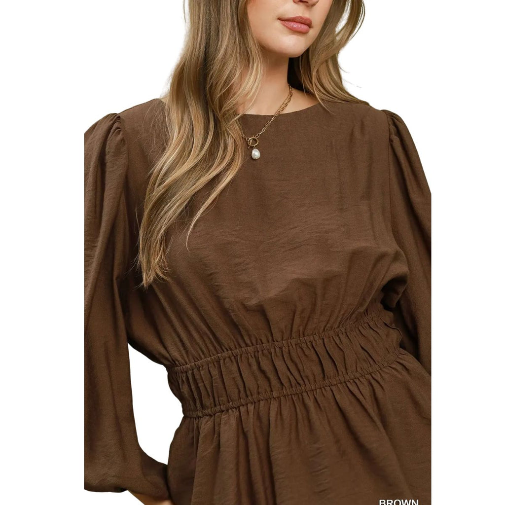 A woman models the Umgee Smocked Waist Long Sleeve Peplum Blouse, featuring puffed shoulders and a smocked elastic waist, shown from shoulders to waist against a white background.