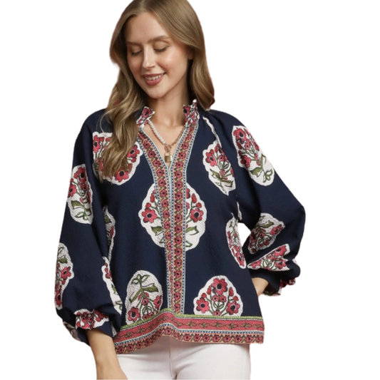 A woman wears the Umgee Boho Floral Top featuring navy blue with red and white flowers, a contrast border, and decorative trim, paired with white pants for a relaxed look.