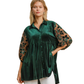 A woman models the Umgee Floral Lace Contrast Velvet Tunic, featuring green velvet with floral lace sleeves and puff details, styled with white pants against a plain background.