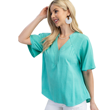 A woman stands against a white background wearing the FASHION GO Washed Linen V-neck Top in turquoise with white pants, accessorized with large white earrings and a necklace.