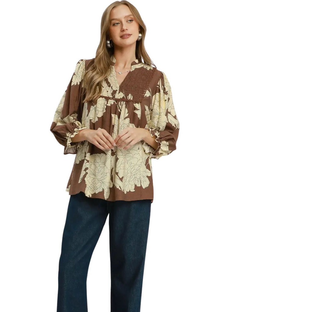 A woman wearing the Umgee Brown and Ivory Floral Peasant Blouse, featuring large cream floral prints, a smocked yoke, and balloon sleeves, paired with dark blue pants and standing against a white background.