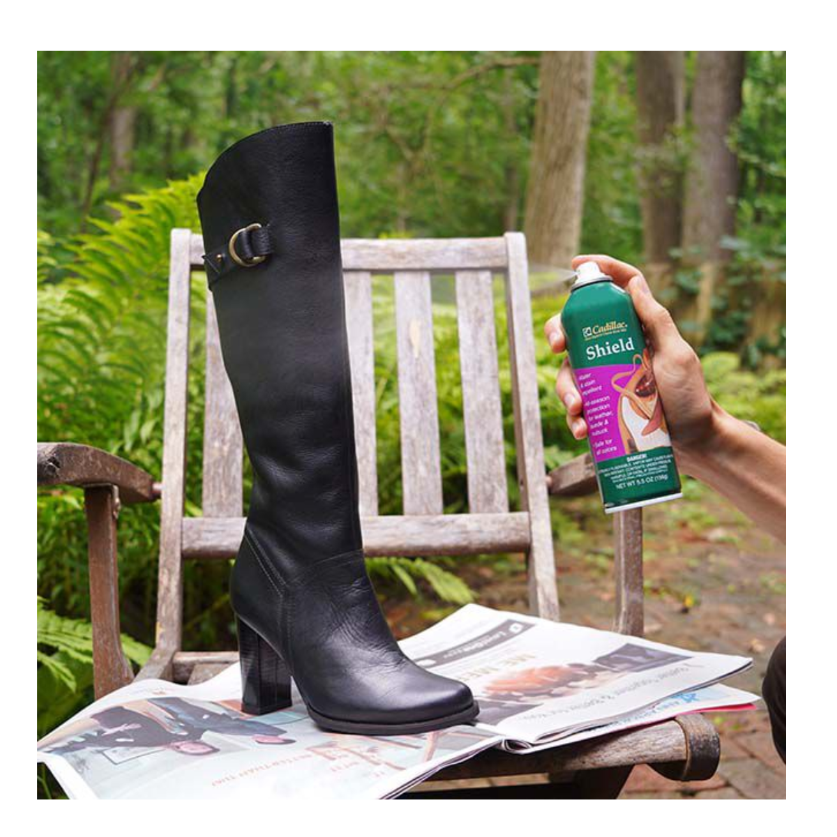 Someone sprays Flipflops & Whatnots Shield Water and Stain Repellent Spray - 5.5oz on a black knee-high boot set on a wooden chair covered with newspapers, outdoors in a garden.