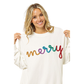 A woman smiles, holding the sides of her FASHION GO Christmas Merry Tinsel Metallic Lettering Pullover, featuring the word "merry" in colorful, textured letters on a white sweater.