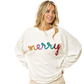 A woman wears the FASHION GO Christmas Merry Tinsel Metallic Lettering Pullover, a white sweater featuring "merry" in colorful, sequined letters, as she stands with her hands on her hips, gazing to the side.