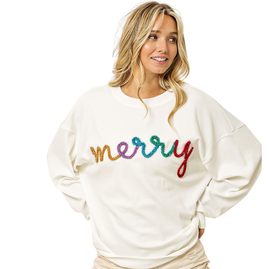 A woman wears the FASHION GO Christmas Merry Tinsel Metallic Lettering Pullover, a white sweater featuring "merry" in colorful, sequined letters, as she stands with her hands on her hips, gazing to the side.