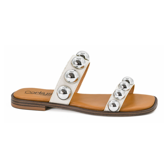 The Catch A Buzz White Flat Sandal by CORKY'S FOOTWEAR INC features two white straps with large silver studs, a tan sole, and a cushioned footbed, shown from the side.