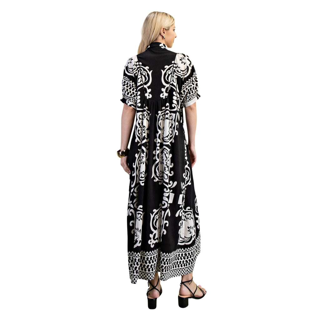 A woman stands with her back to the camera, wearing the FASHION GO Deserving Black and White Print Dress featuring a smocked yoke and bold mixed print patterns, paired with black sandals, earrings, and a bracelet, set against a white background.