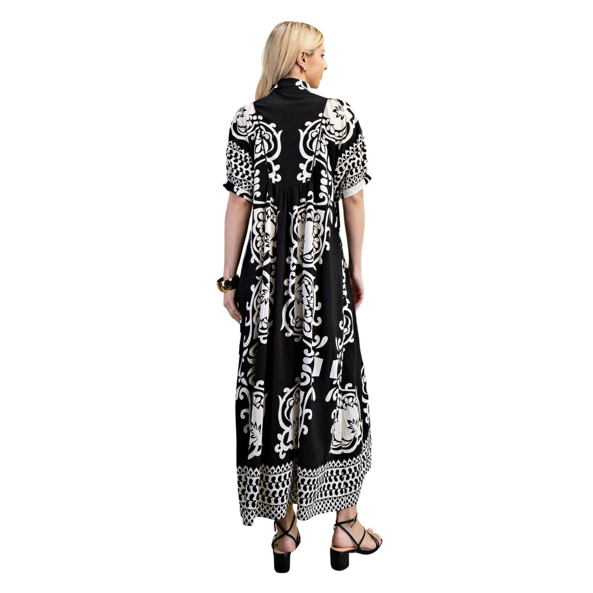 A woman stands with her back to the camera, wearing the FASHION GO Deserving Black and White Print Dress featuring a smocked yoke and bold mixed print patterns, paired with black sandals, earrings, and a bracelet, set against a white background.