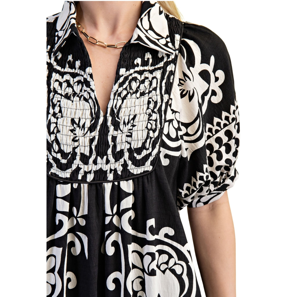 A person wearing the Deserving Black and White Print Dress by FASHION GO, featuring white floral patterns, a deep V-neck, puffed sleeves, and accessorized with a gold chain necklace.