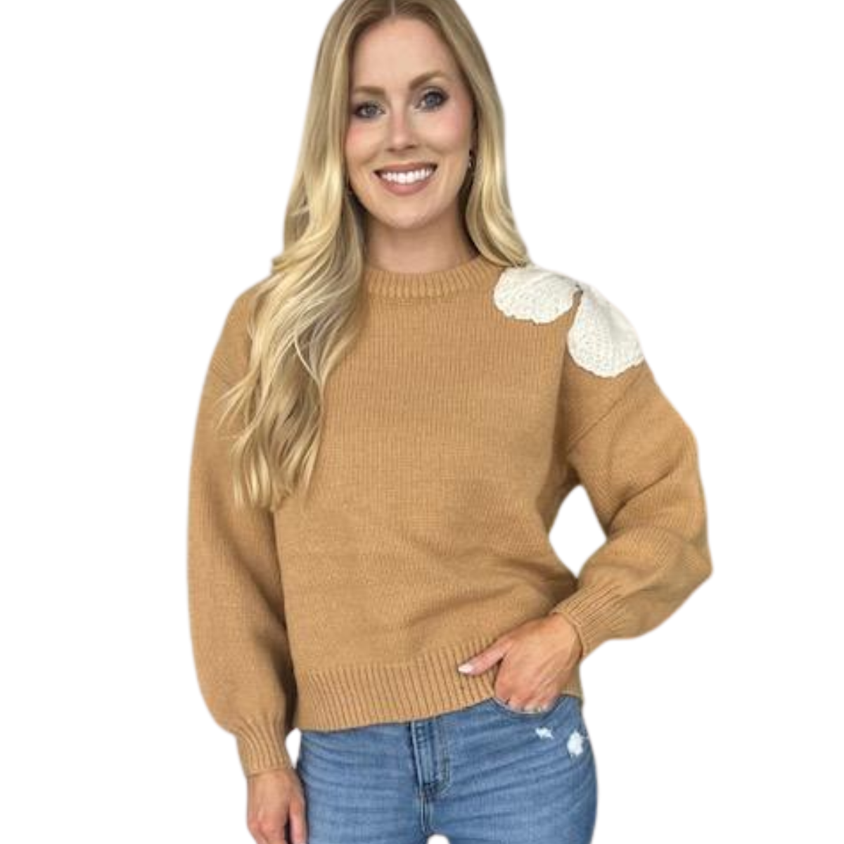 A woman wears the Carole Christian Bow Shoulder Sweater in Camel, featuring an ivory flower bow accent on the left shoulder, paired with blue jeans against a white background.
