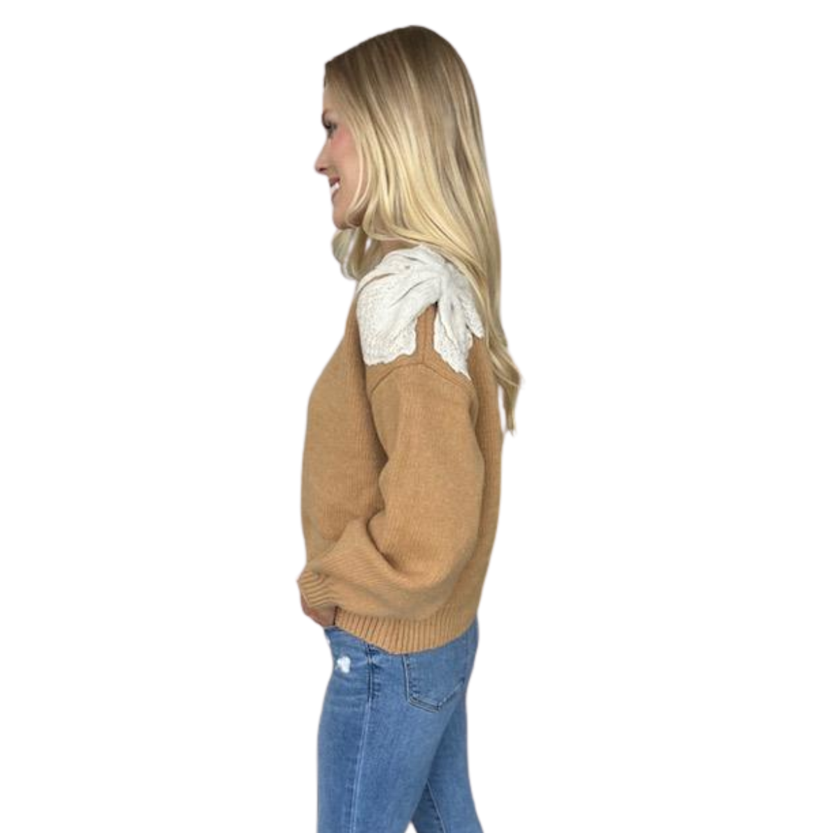 A woman stands in profile against a plain white background wearing the Carole Christian Bow Shoulder Sweater in Camel with white lace detail on the shoulder, paired with blue jeans.