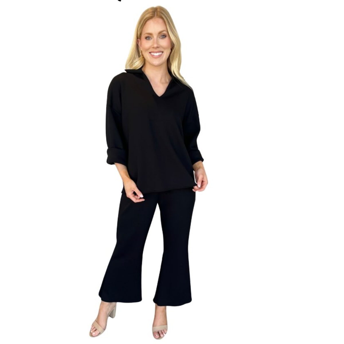 A woman stands smiling in the Carole Christian Modal Scuba Fabric Set—an elegant black two-piece with three-quarter sleeves and wide-leg pants, styled with light heels for seamless day-to-night wear against a white background.