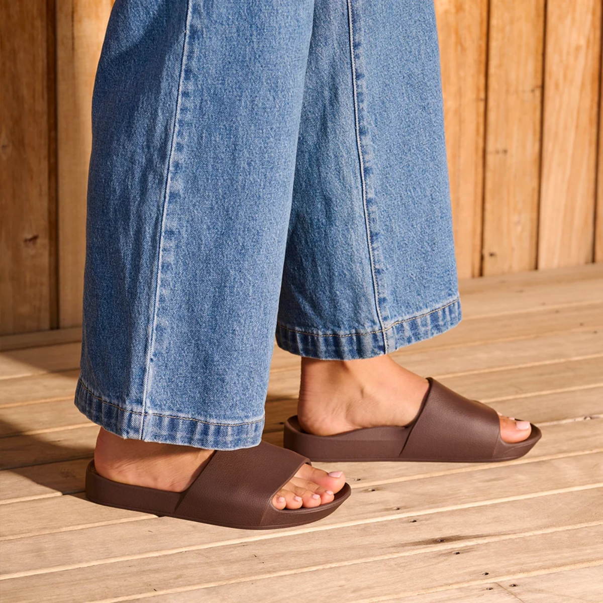 Wearing wide-leg blue jeans and Archies Slide Sandal in Brown by ARCHIES FOOTWEAR LLC, a person stands on a wooden deck in front of a wooden wall.