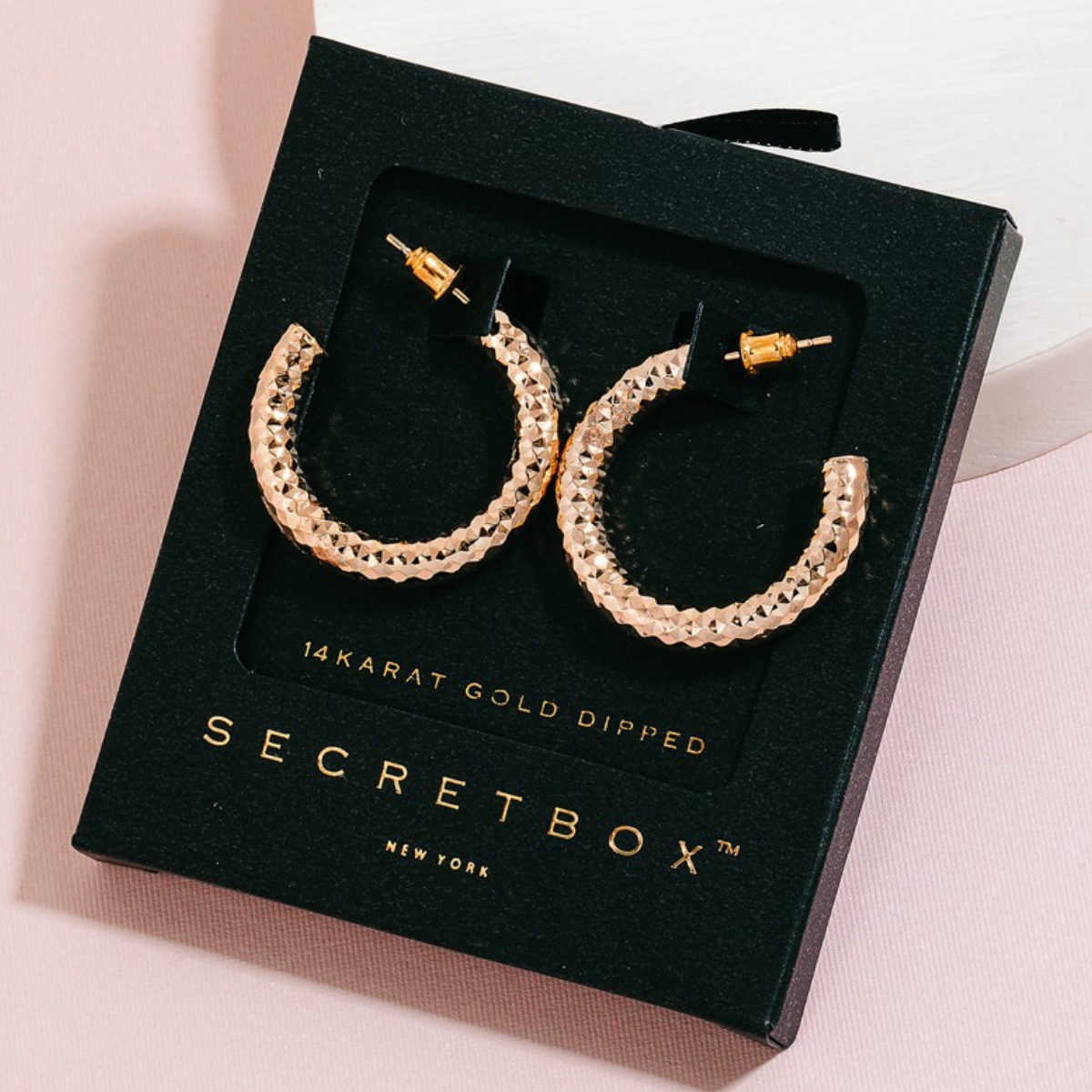 Pair of Secret Box Gold Dipped Textured Hoop Earrings in Gold by FASHION GO in a black box labeled "SECRET BOX NEW YORK," displayed against a pink background. Main image